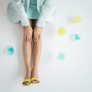 From above crop unrecognizable ethnic female lying on white floor in light blue apparel and yellow shoes with transparent glasses with yellow and blue liquids