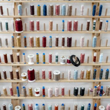 Stand with assorted colorful spools of thread for sewing placed near white wall inside dressmaker workshop or tailor atelier for fashion design and apparel creation